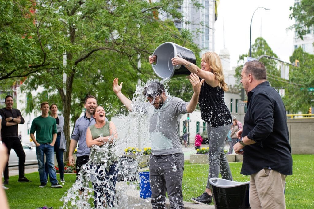 Das Bild zeigt eine Gruppe von Menschen, die an der Ice Bucket Challenge teilnehmen. In der Mitte ist eine Frau zu sehen, die einen Eimer mit Wasser über den Kopf eines Mannes schüttet.