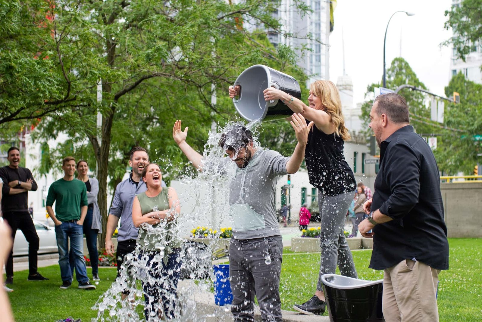 Das Bild zeigt eine Gruppe von Menschen, die an der Ice Bucket Challenge teilnehmen. In der Mitte ist eine Frau zu sehen, die einen Eimer mit Wasser über den Kopf eines Mannes schüttet.