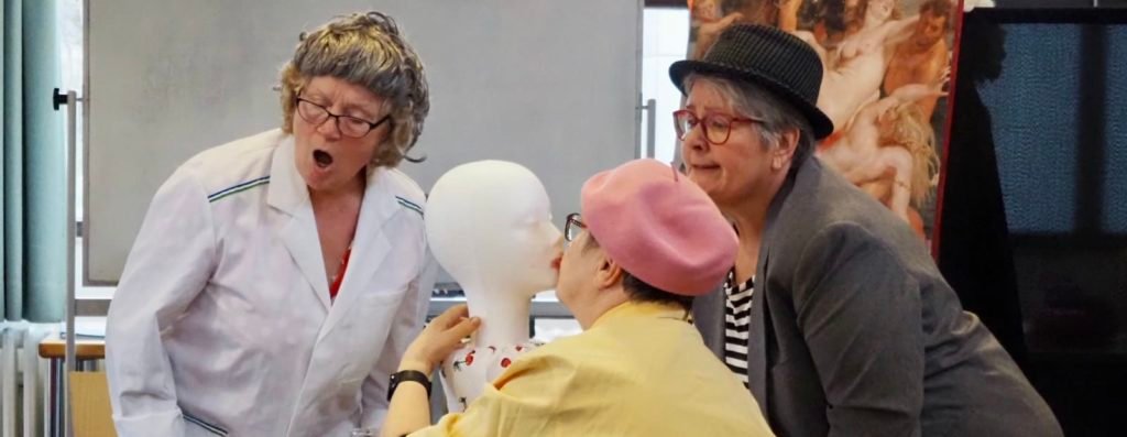 Drei Schauspielerinnen, eine von ihnen küsst einen Kopf aus Styropor