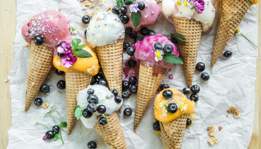 Mehrere Waffeltüten mit unterschiedlichen Eissorten liegen auf einem Tisch. Auf ihnen sind Blaubeeren und Glitzer verstreut.