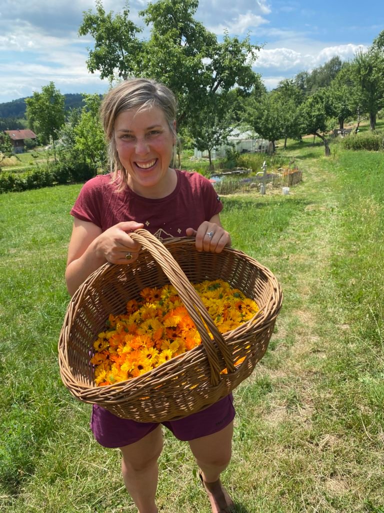 Eine Frau hält einen Korb in der Hand, darin viele gelbe Blüten, sie steht draußen auf einem Feld und lacht, WWOOF
