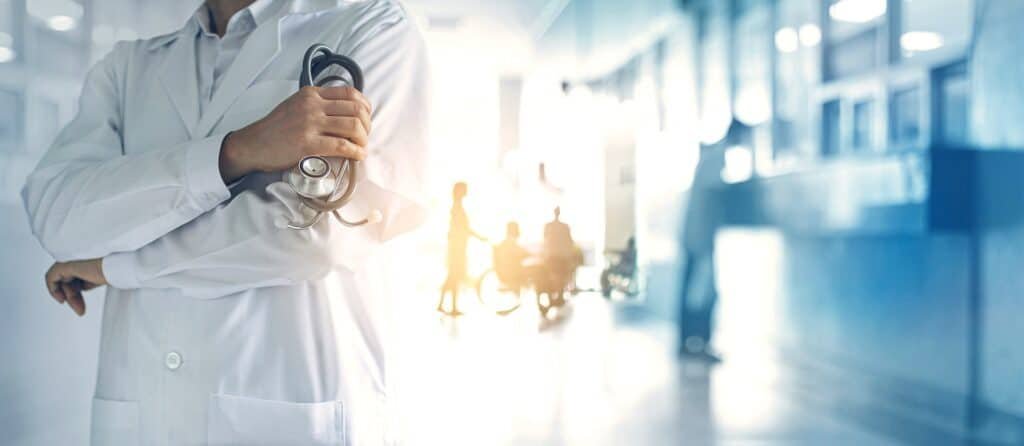 Vordergrund: Person in weißem Arztkittel hält ein Stethoskop. Im Hintergrund sind unscharf Patient:innen zu erkennen, die Multiple Sklerose haben könnten.