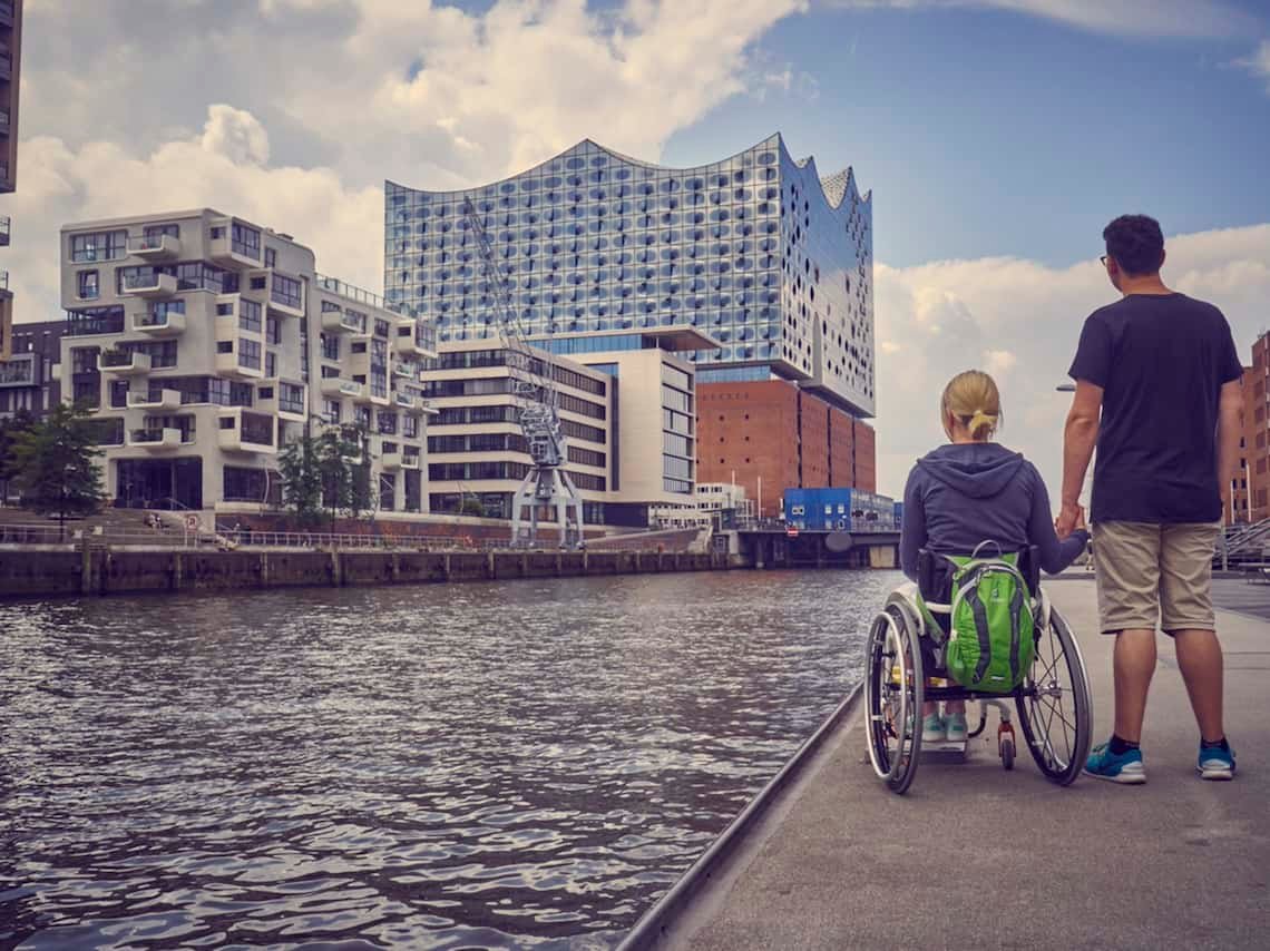 Zu sehen ist das Hamburger Konzerthaus "Elbphilharmonie" vor einem hellblauen Himmel mit großen, weißen Wolken. Vor der Elbphilharmonie fließt die Elbe. Am gegenüberliegenden Ufer stehen eine Rollstuhlfahrerin und ein Mann, sie halten sich an der Hand und schauen auf die Elbphilharmonie.