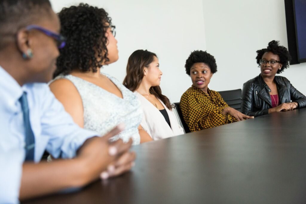 Fünf Personen unterschiedlichen Geschlechts und mit vielfältigem Erscheinungsbild sitzen an einem dunklen Konferenztisch und sind in ein Gespräch vertieft. Sie sind in einer modernen Umgebung, vermutlich in einem Konferenzraum. Eine Person in der Mitte trägt ein gemustertes Oberteil und spricht, während die anderen aufmerksam zuhören. Die Stimmung wirkt offen und kommunikativ, was auf eine Diskussion oder einen Austausch in einem multikulturellen Team hinweist. Die Gruppe spiegelt Diversität und Zusammenarbeit wider.