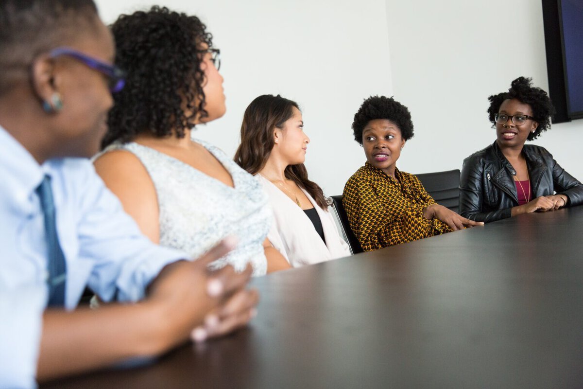 Fünf Personen unterschiedlichen Geschlechts und mit vielfältigem Erscheinungsbild sitzen an einem dunklen Konferenztisch und sind in ein Gespräch vertieft. Sie sind in einer modernen Umgebung, vermutlich in einem Konferenzraum. Eine Person in der Mitte trägt ein gemustertes Oberteil und spricht, während die anderen aufmerksam zuhören. Die Stimmung wirkt offen und kommunikativ, was auf eine Diskussion oder einen Austausch in einem multikulturellen Team hinweist. Die Gruppe spiegelt Diversität und Zusammenarbeit wider.