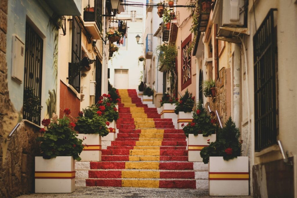 Schmale, von weißen Häusern gesäumte Gasse mit einer Treppe in Rot und Gelb, flankiert von Blumenkästen mit roten Geranien und kleinen Balkonen.