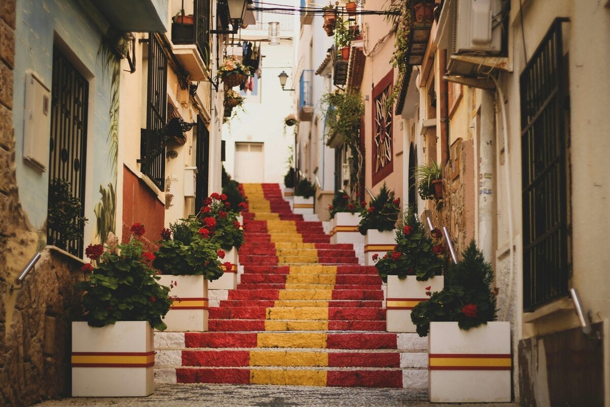 Schmale, von weißen Häusern gesäumte Gasse mit einer Treppe in Rot und Gelb, flankiert von Blumenkästen mit roten Geranien und kleinen Balkonen.